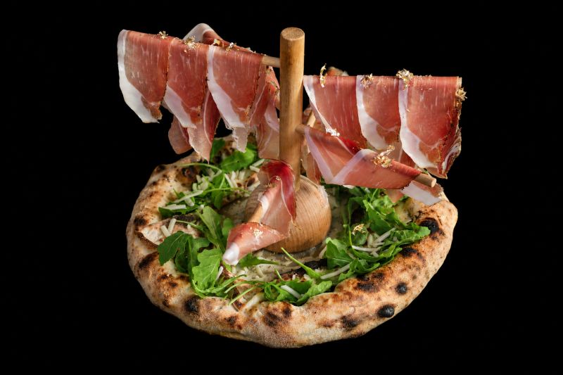 La pizza Life con la cascata di speck appesa a un supporto di legno, rucola e scaglie d'oro